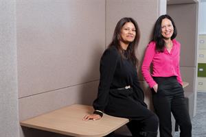 Kulsoom Ali (left) and Suzy McCormick both sit on a wooden desk in front of a beige wall