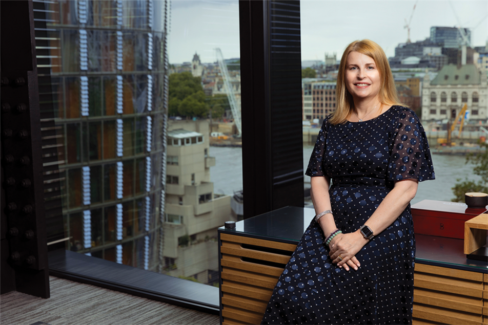Julia Paulding is wearing a navy dress and leaning on a desk. Behind her is a full-length window with a view of the River Thames in central London