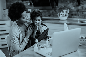 Woman with child on her lap holding a tissue to his nose in front of laptop